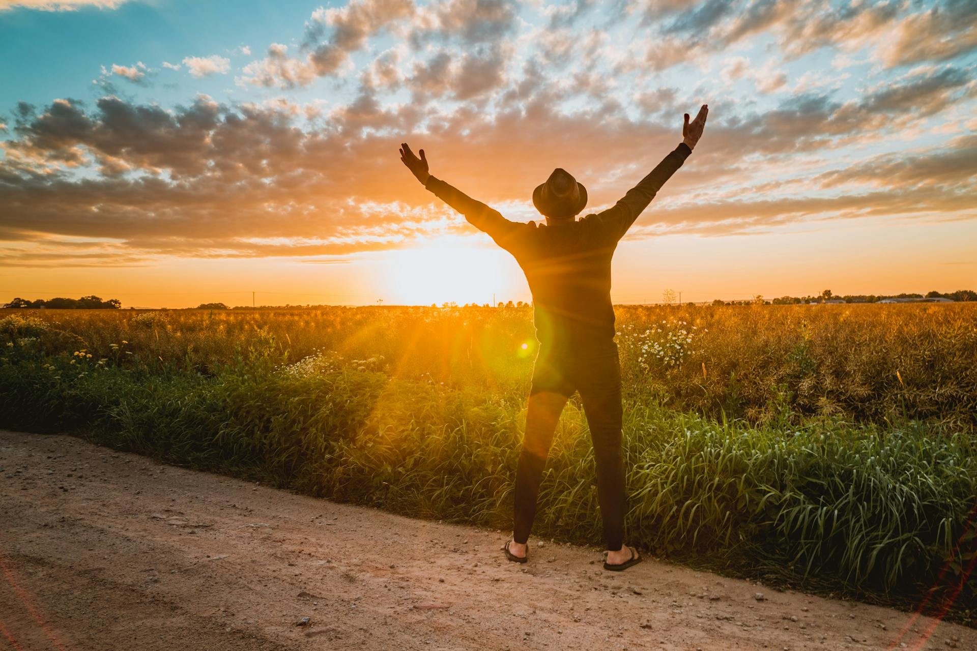 A man raises arms in freedom at sunrise in a rural field, expressing joy and connection with nature.