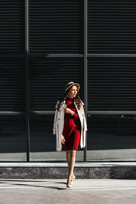 Full body trendy female with long curly hair in red dress and hat and high heeled shoes and coat adjusting collar while walking along road from contemporary building in daylight
