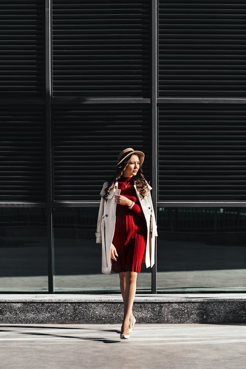 Full body trendy female with long curly hair in red dress and hat and high heeled shoes and coat adjusting collar while walking along road from contemporary building in daylight