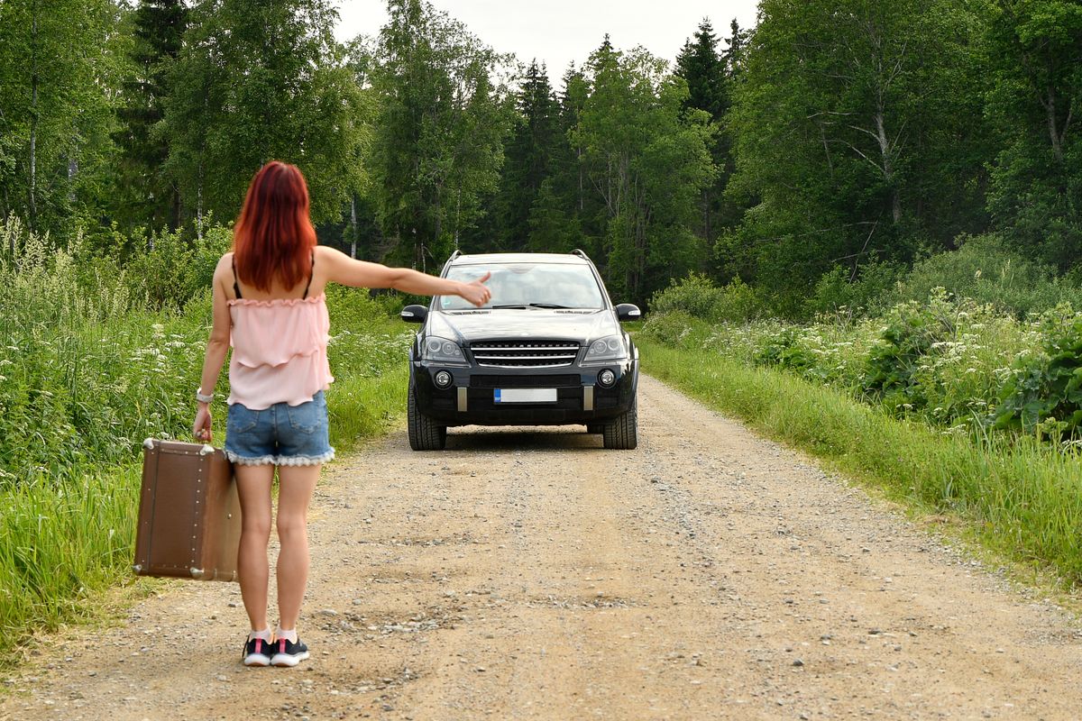 Autostopista cu degetul ridicat si o masina oprita in fata ei pe un drum forestier