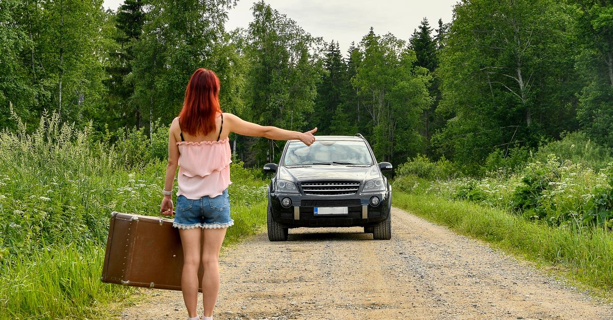 A woman with red hair hitchhikes on a rural road in Kolga, Estonia, holding a vintage suitcase.