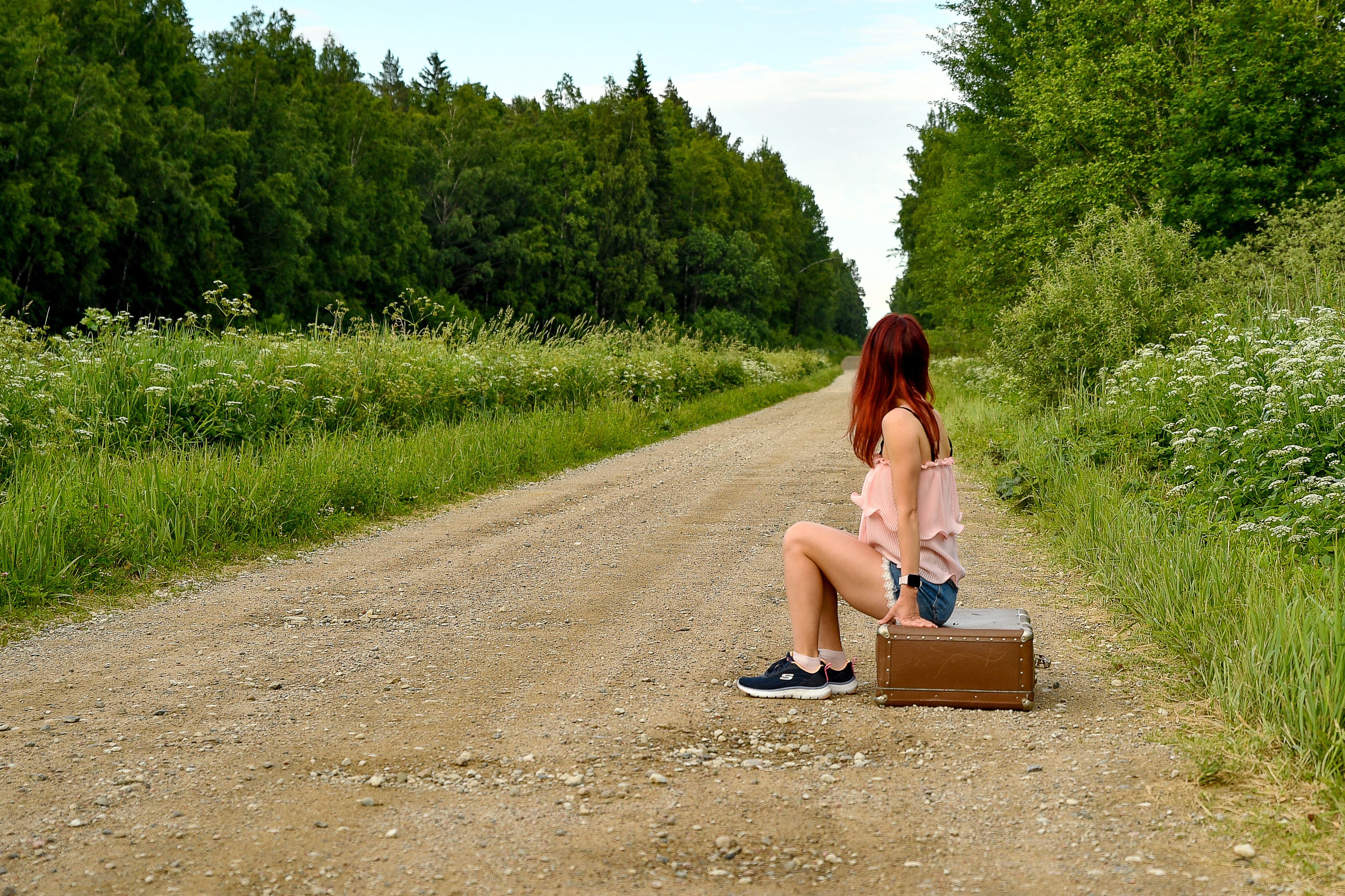 A woman with red hair sits on a suitcase on a gravel road in a lush green forest setting.