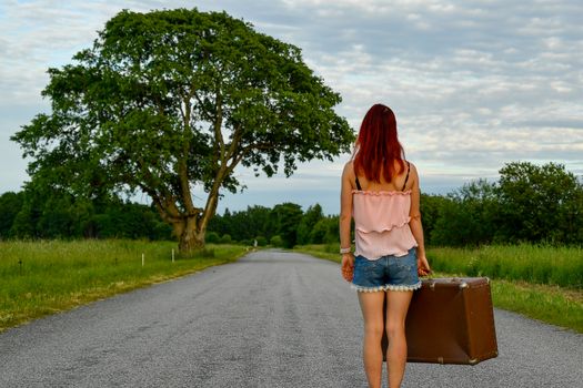 Woman in summer attire walking along a country road with vintage luggage, symbolizing travel and exploration.