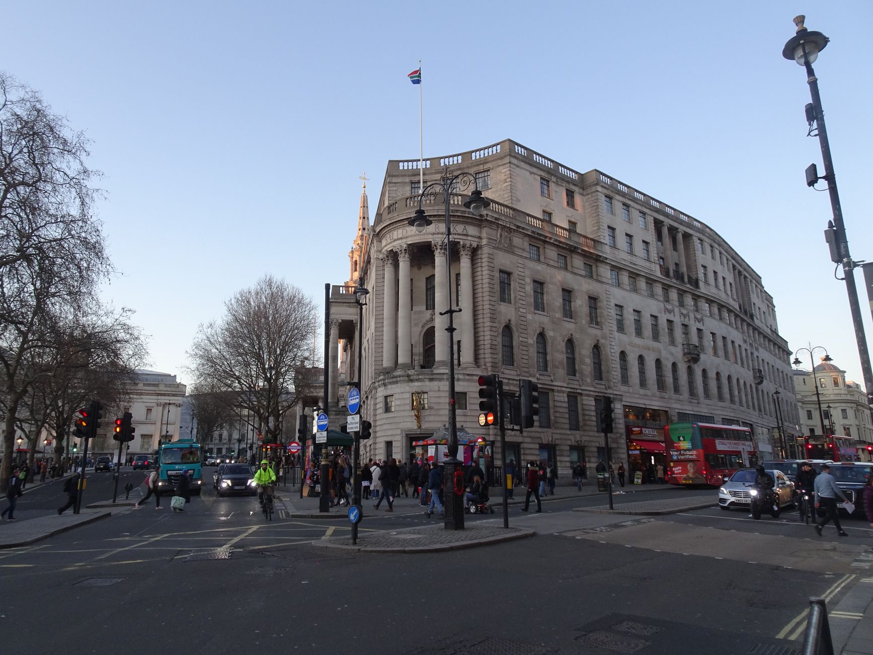 Busy street in London with classic architecture, buses, and pedestrians under a blue sky.