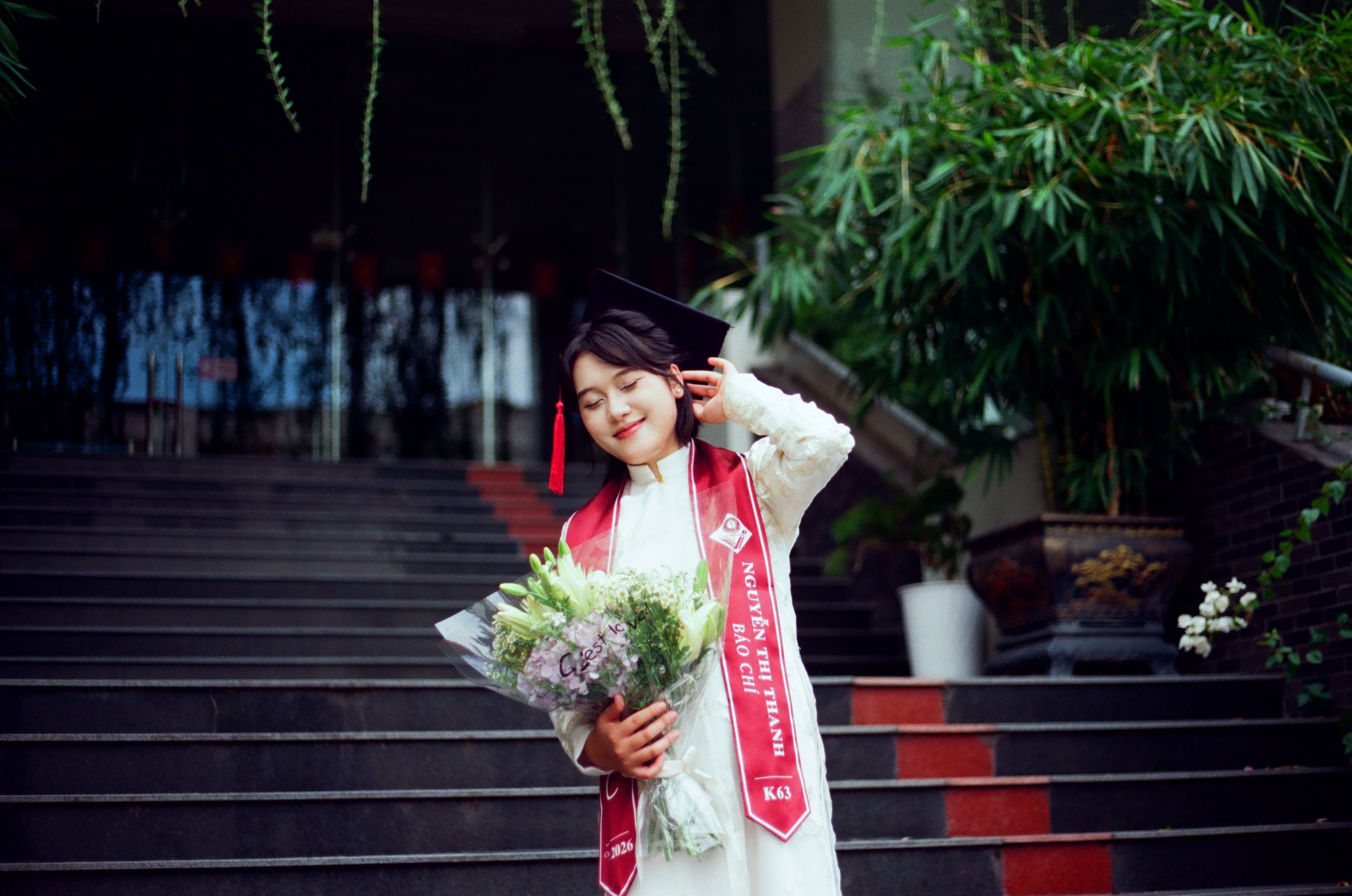 A joyful graduate wearing a cap and gown poses with a bouquet on an outdoor staircase.