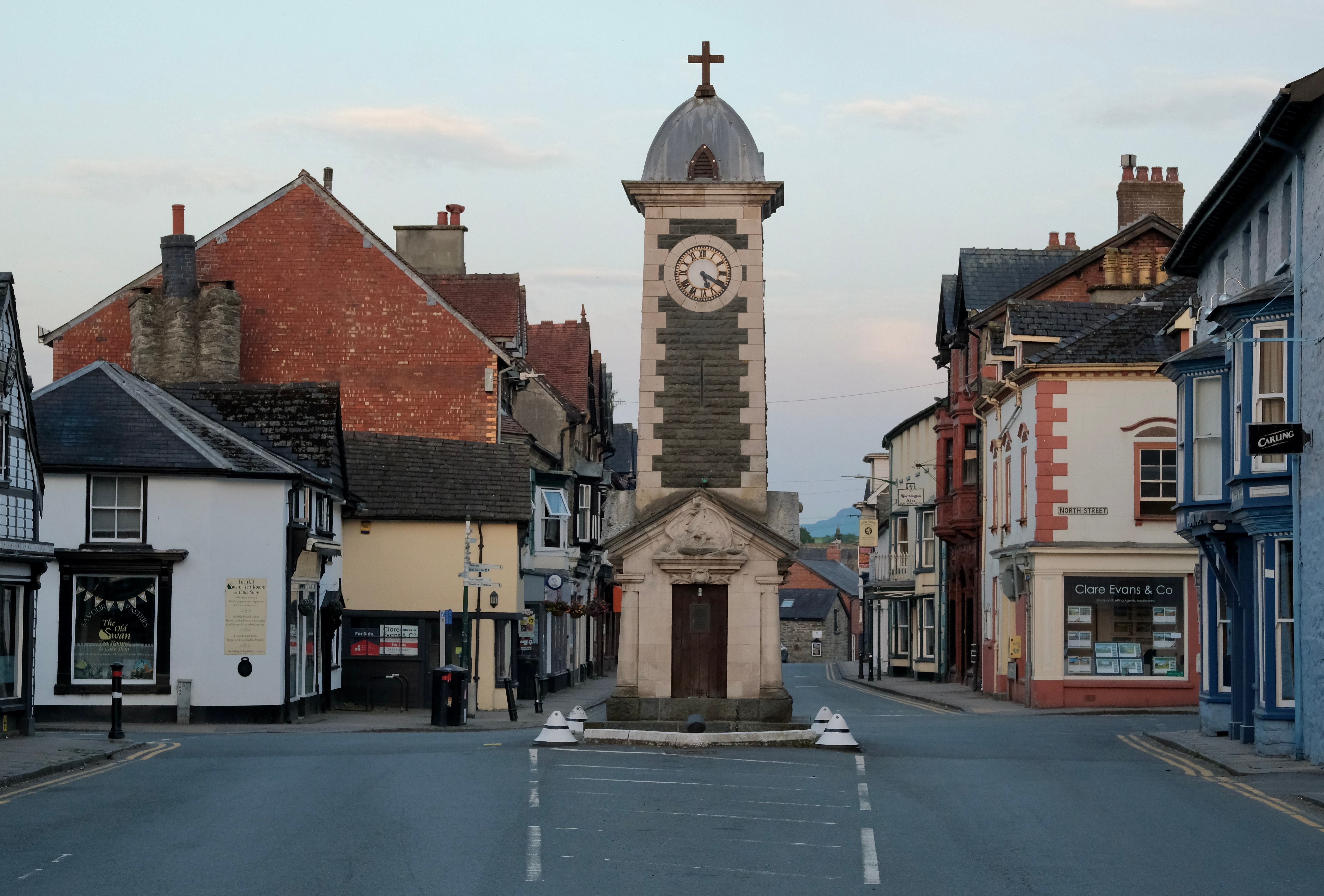 View of a historic town square in the UK featuring a central stone clock tower and traditional buildings.