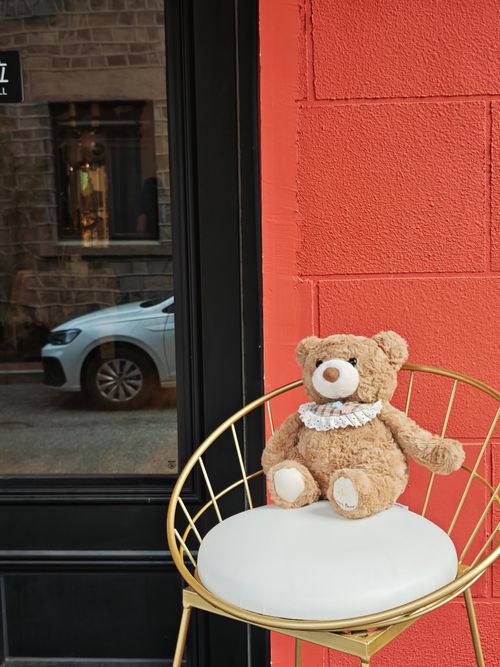 Free Cozy teddy bear sits elegantly on a gold chair outside a shop with a vibrant orange wall. Stock Photo
