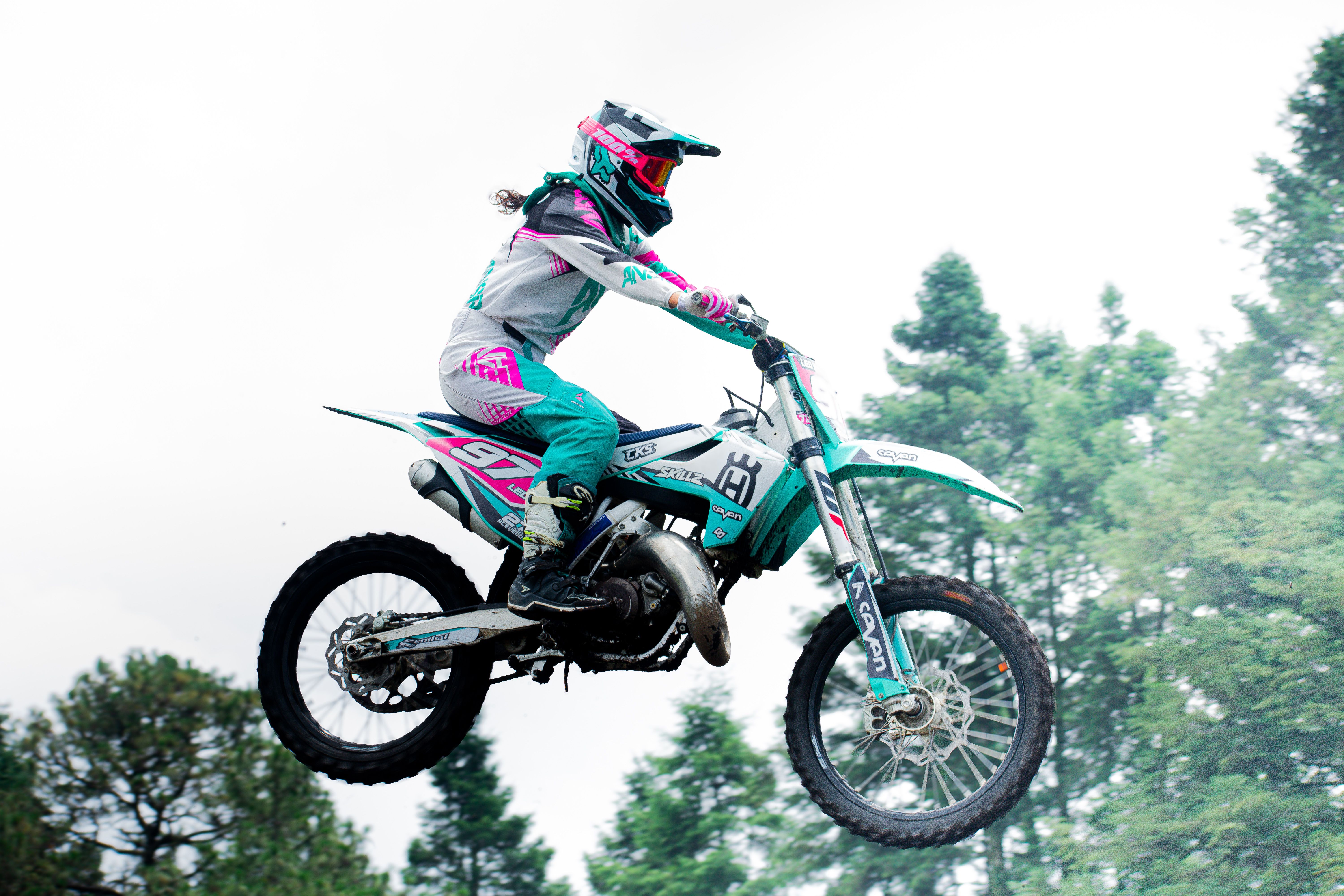 A motocross rider in vibrant gear performs a high jump against a forest backdrop in Mexico City.