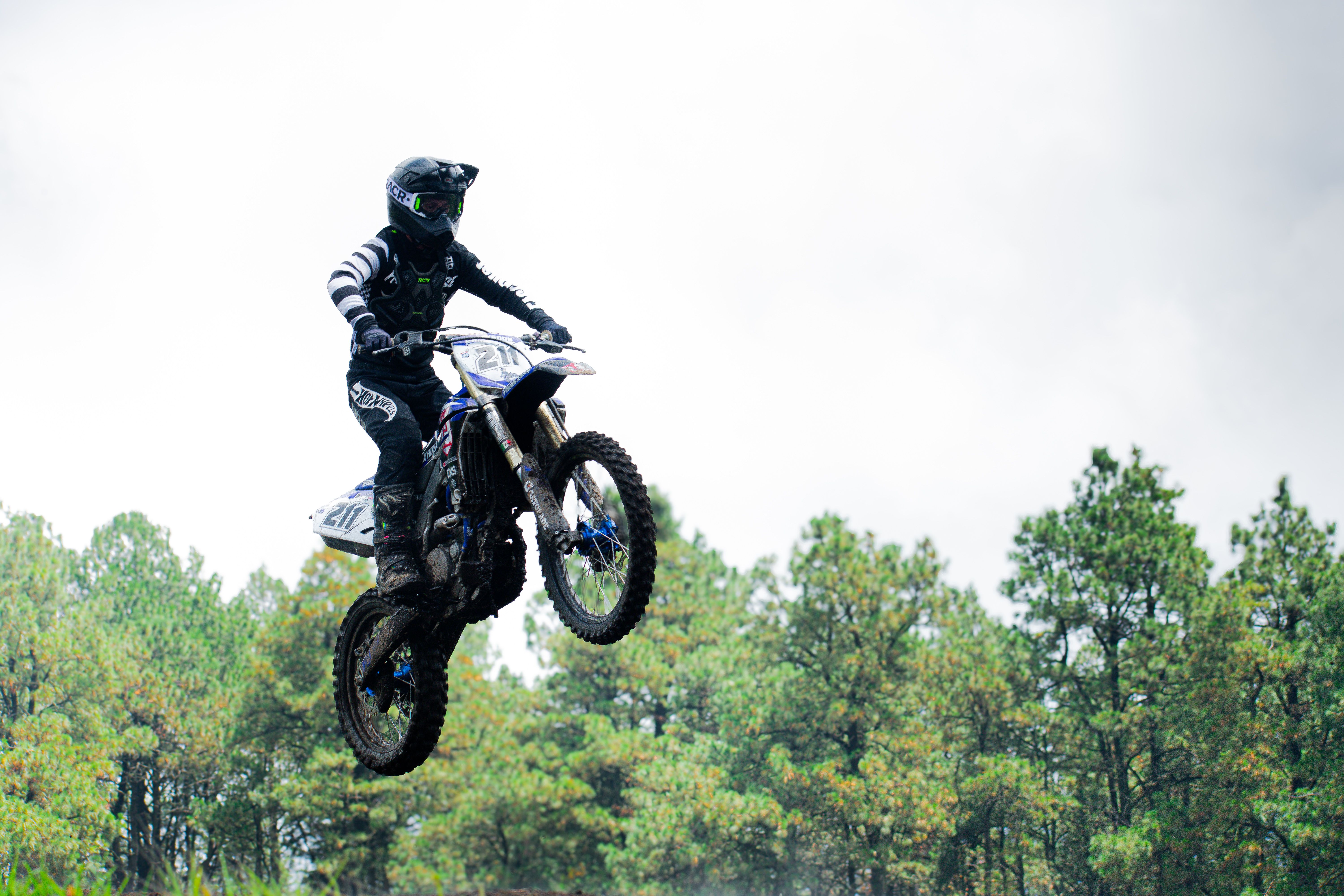 A motocross rider performs a high jump against a scenic forest background.
