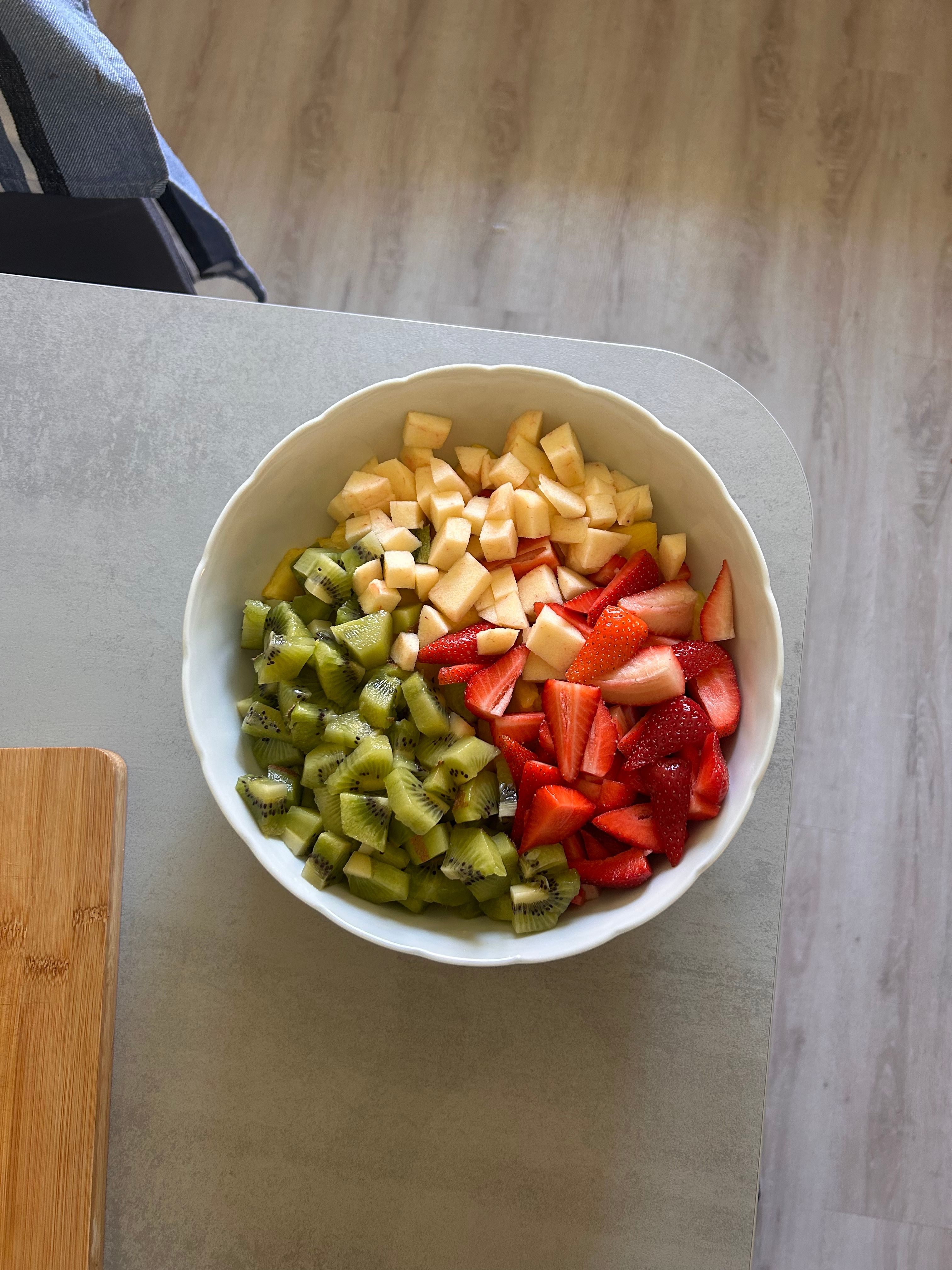 A top view of a vibrant fruit salad in a white bowl with kiwi, apple, and strawberry.