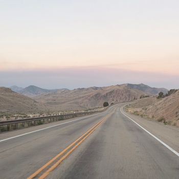 A winding desert highway stretches into distant mountains under a pastel sunset sky.
