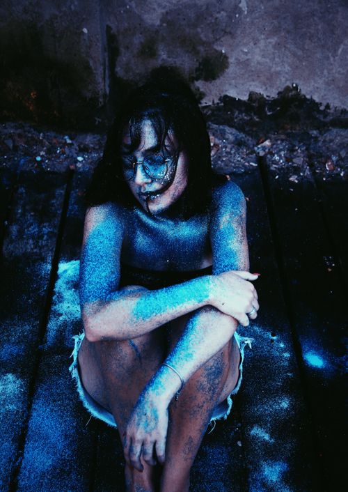 Free From above calm young fit female wearing casual outfit with skin covered with blue fluorescent paint sitting on floor in studio and hugging knees Stock Photo
