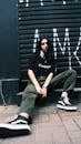 Pretentious young female wearing casual street style clothing sitting relaxed on sidewalk pavement against grungy wall and looking at camera