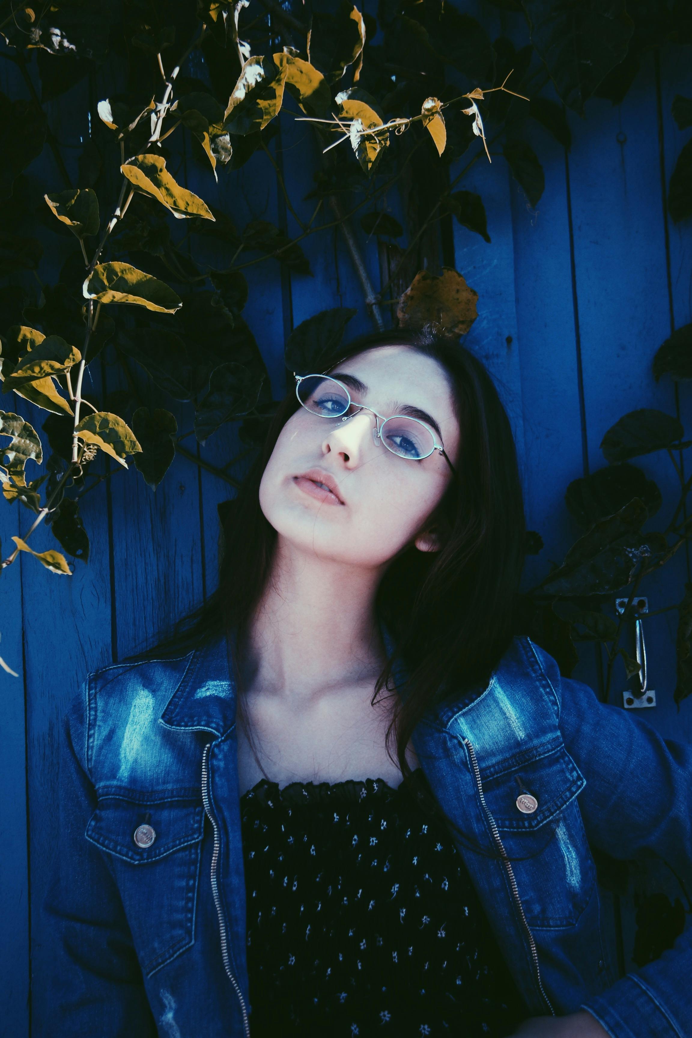 Portrait of a woman in glasses and denim jacket against a blue wooden backdrop with foliage.