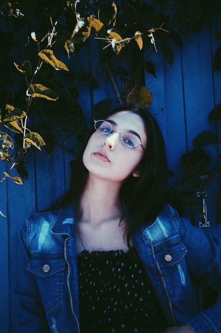 Portrait of a woman in glasses and denim jacket against a blue wooden backdrop with foliage.