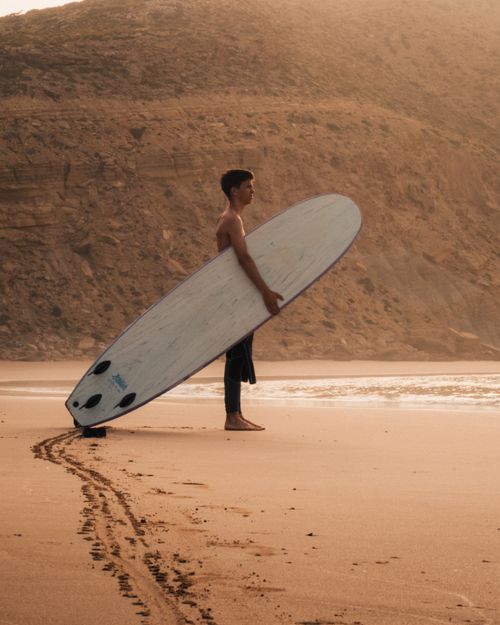 Free A surfer stands with a board on a sandy beach at sunset, creating a peaceful seaside atmosphere. Stock Photo