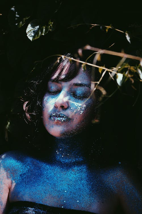 Relaxed female model with eyes closed standing with blue glittering paint on skin and dramatic makeup in studio near plant twigs