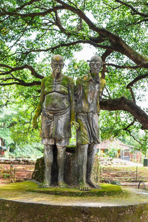 Free Moss-covered stone statue of elderly couple in outdoor park surrounded by verdant trees. Stock Photo