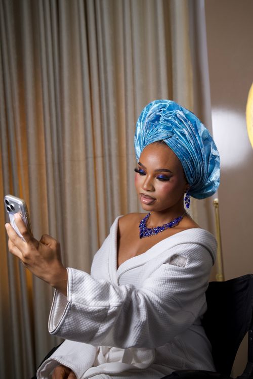 Free A woman in a blue gele headwrap and robe takes a selfie indoors, showcasing cultural elegance and beauty. Stock Photo