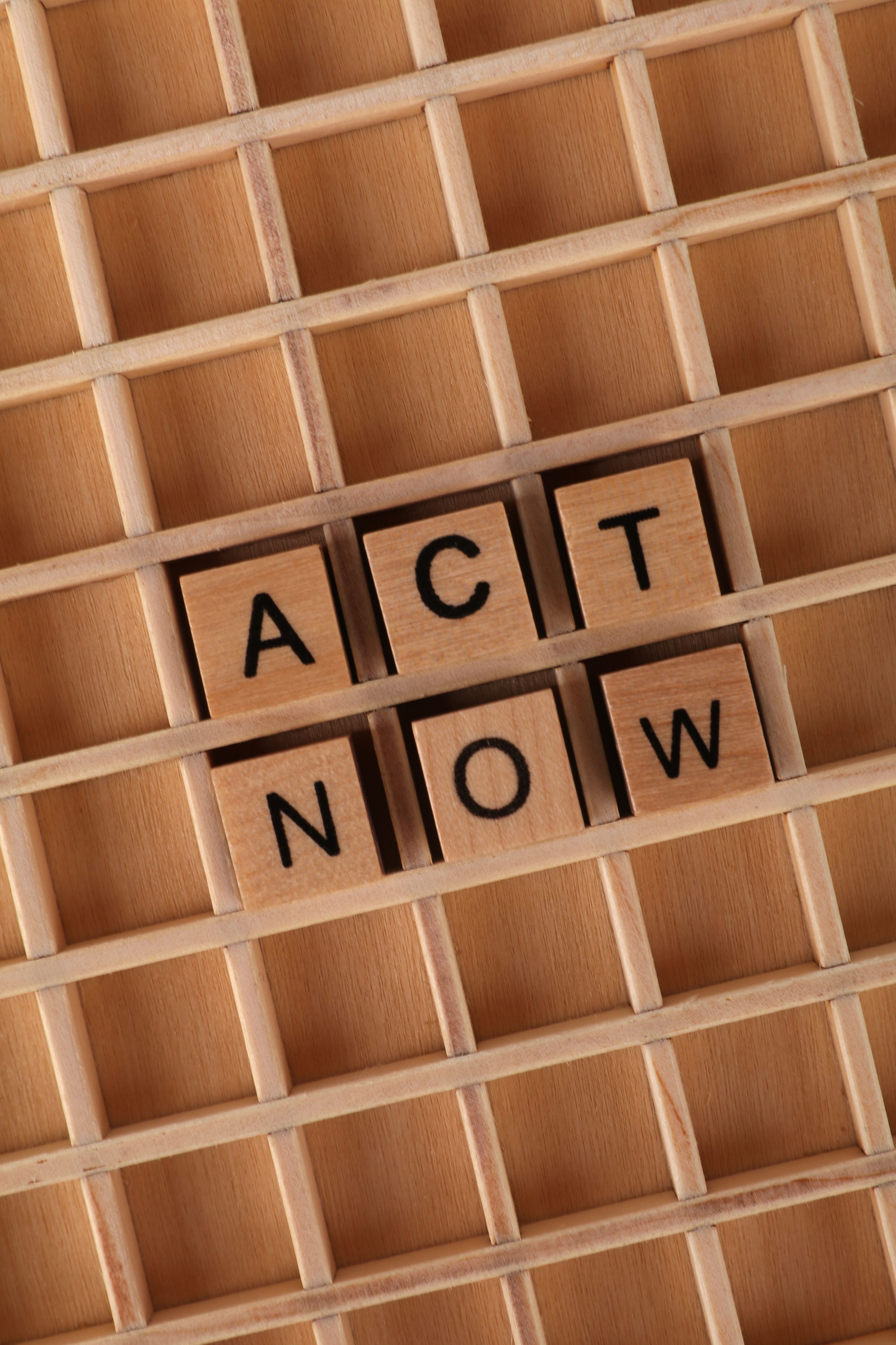 Wooden Scrabble tiles spelling 'Act Now' on a wooden grid, urging immediacy.