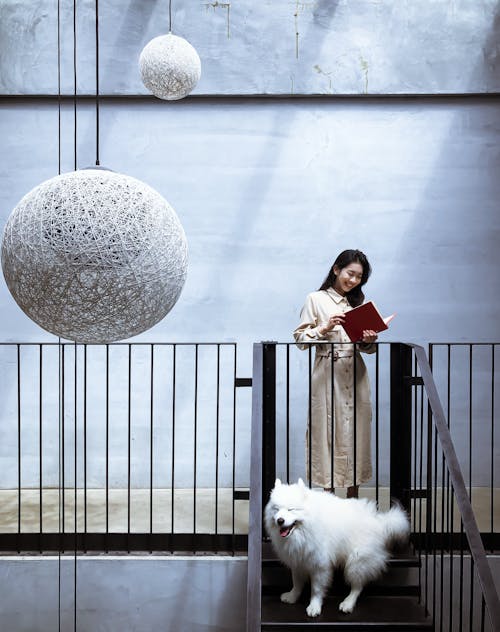 Woman in Beige Shirt Dress Holding a Book Beside White Furry Dog