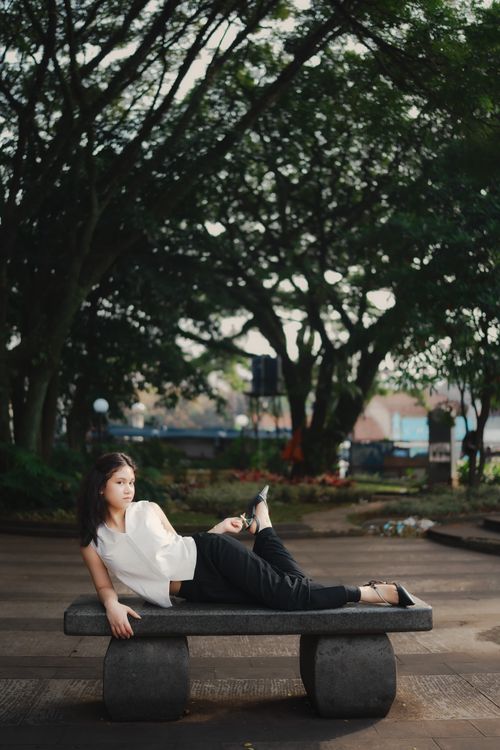Free Young woman relaxing on a stone bench under lush trees in a Bandung park. Stock Photo