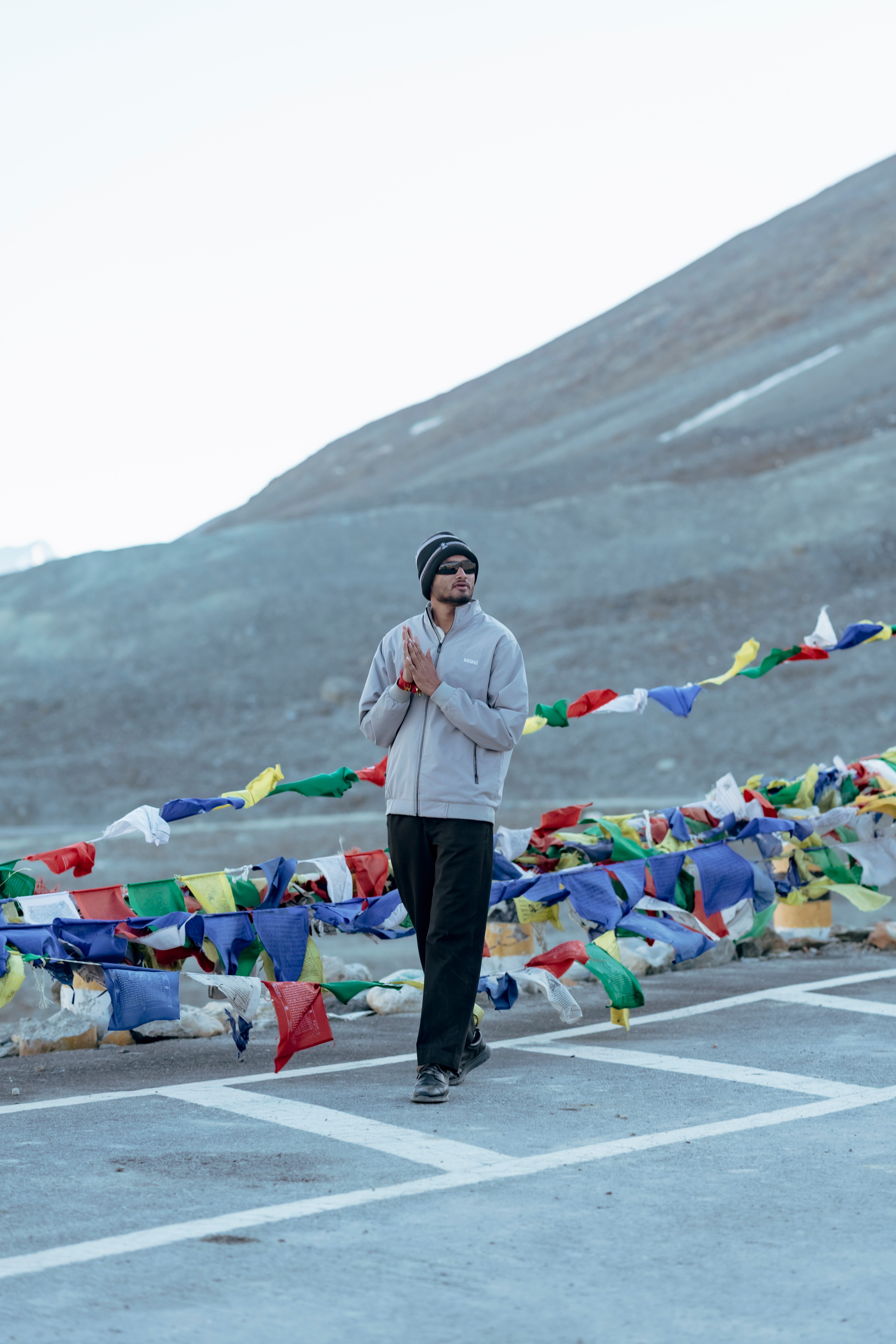A man stands by colorful prayer flags in a serene mountainous setting under a clear sky.