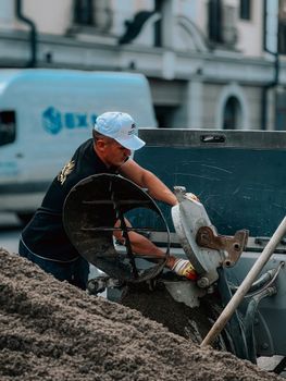 Male construction worker wearing a cap operates a cement mixer on a city street, showcasing urban development.