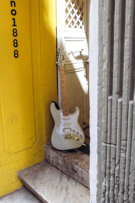 A white electric guitar rests on steps in a sunlit alley, framed by vibrant yellow walls.