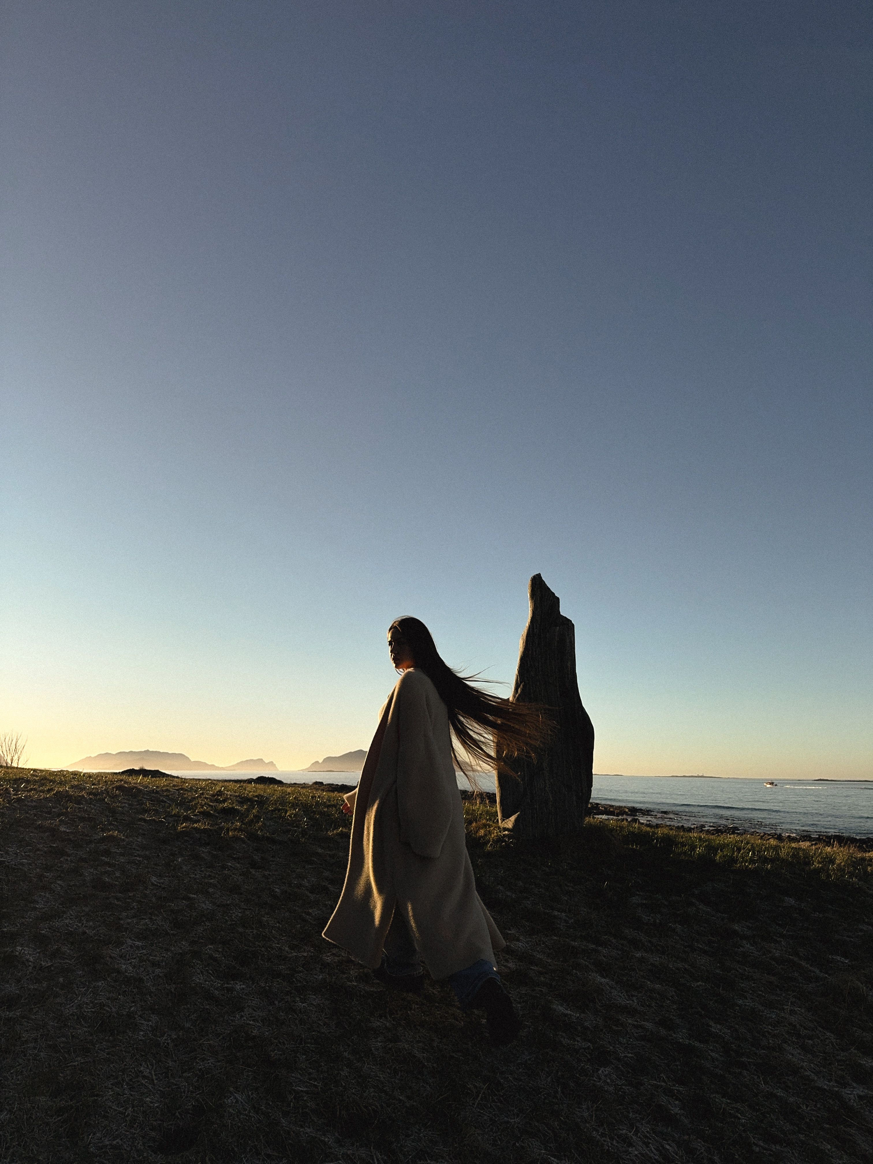 A woman walks past a stone structure at sunset along the Norwegian coast, capturing a serene silhouette.