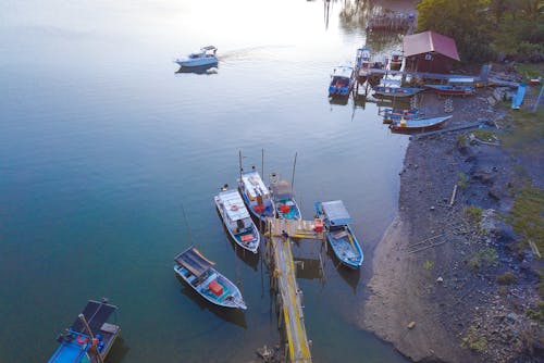From above boats moored near wooden weathered pier and cozy small cottage on waterfront in rural area