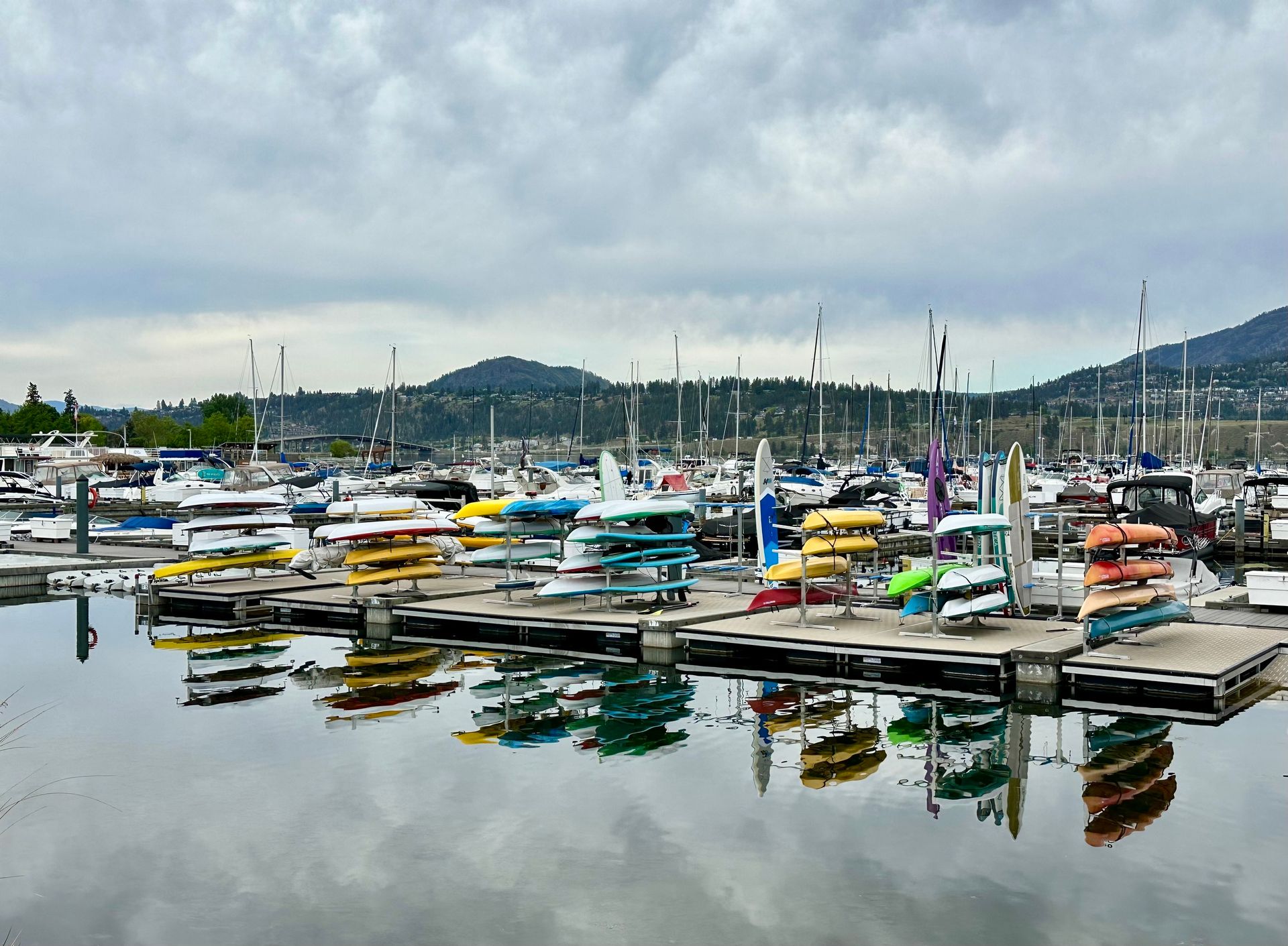 Vibrant kayaks and boats reflected in the calm waters of a marina in Kelowna, with a mountainous backdrop.