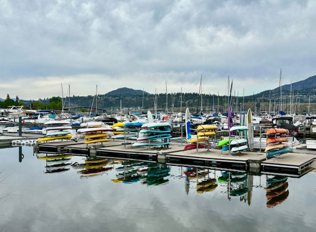 Vibrant kayaks and boats reflected in the calm waters of a marina in Kelowna, with a mountainous backdrop.