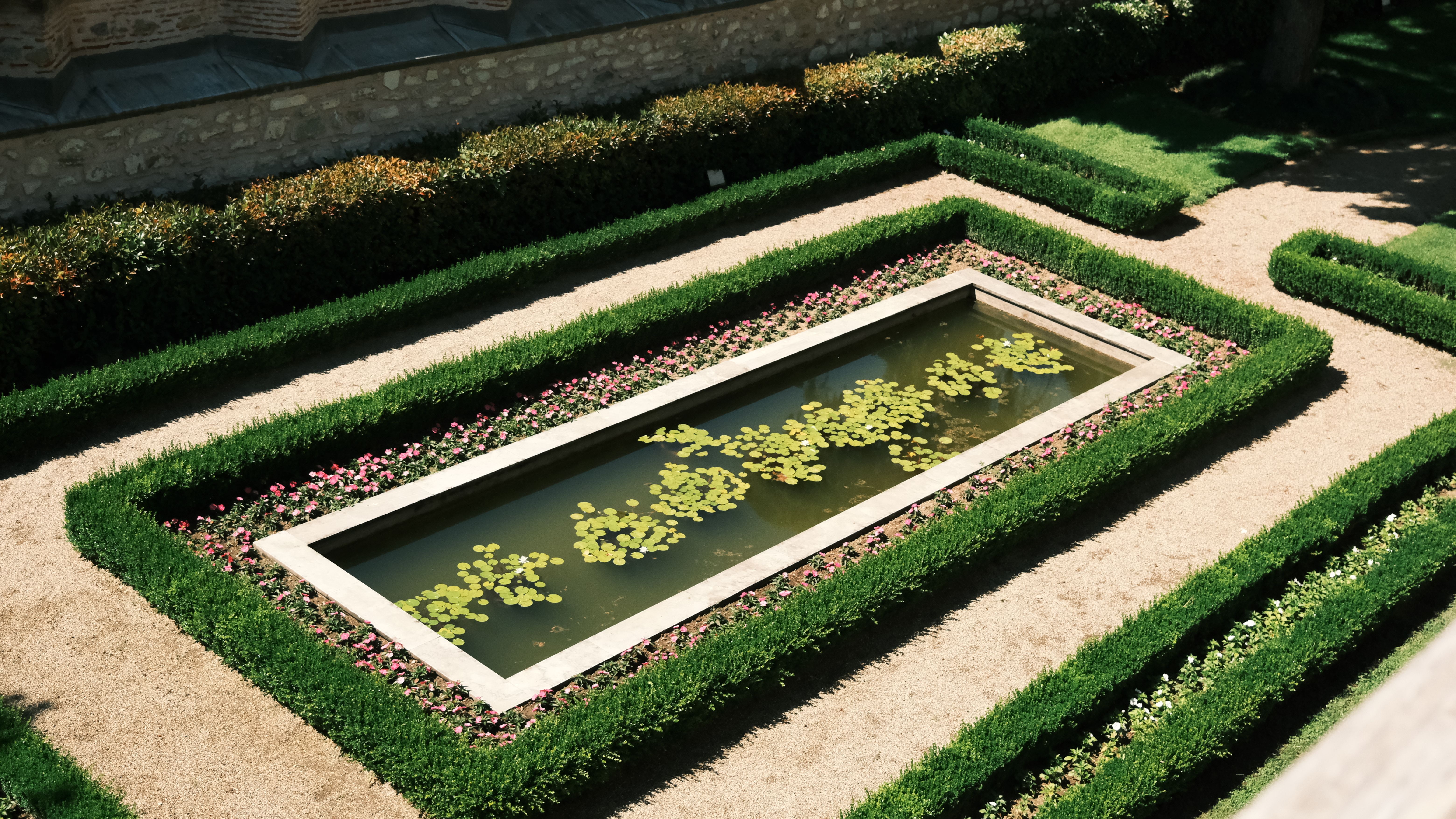 A top view of an elegant rectangular pond surrounded by symmetrical hedges and flowers, in bright daylight.