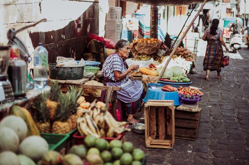 Free Elderly vendor in a bustling street market surrounded by fresh fruits and vegetables on a sunny day. Stock Photo
