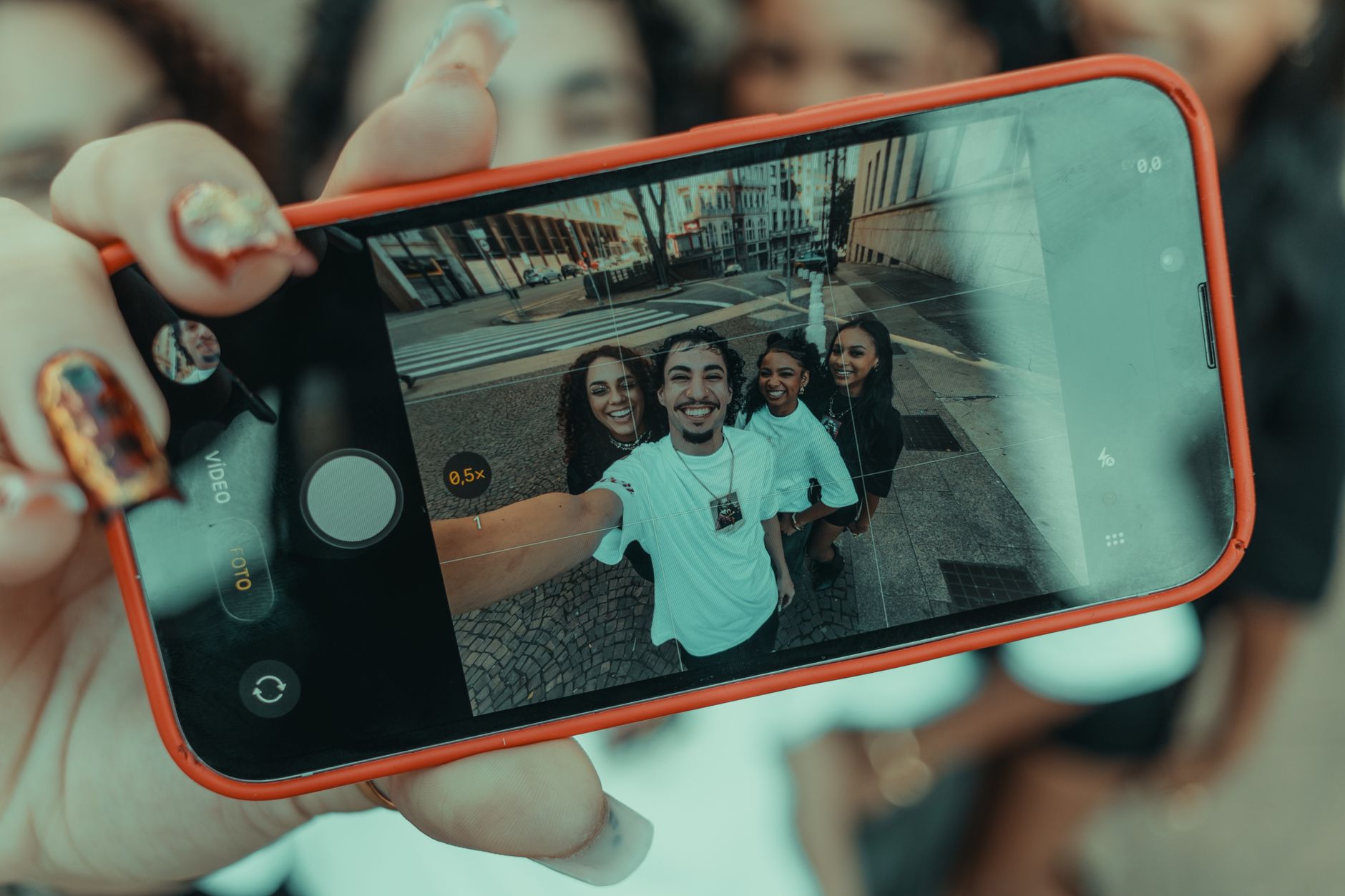 Main tenant un smartphone à coque orange affichant à l'écran un groupe de jeunes souriants en selfie
