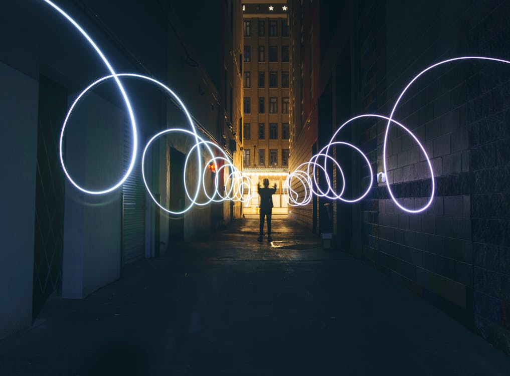 Long exposure full body person silhouette making circles with bright flashlight while standing on narrow dark city street between tall urban buildings