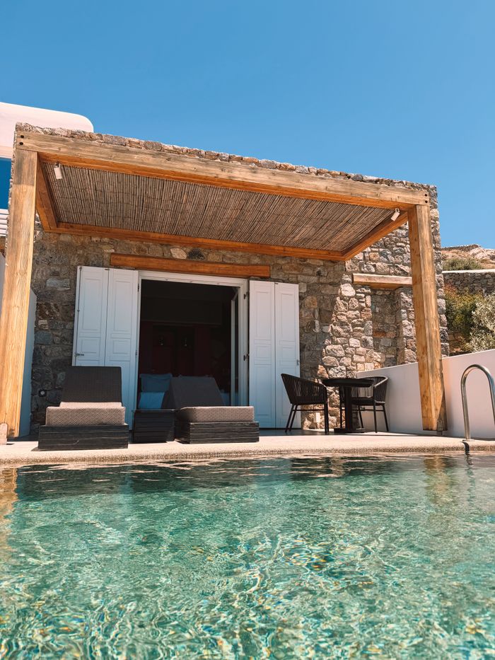 A small stone house with white shutters under a reed pergola, a clear plunge pool in front