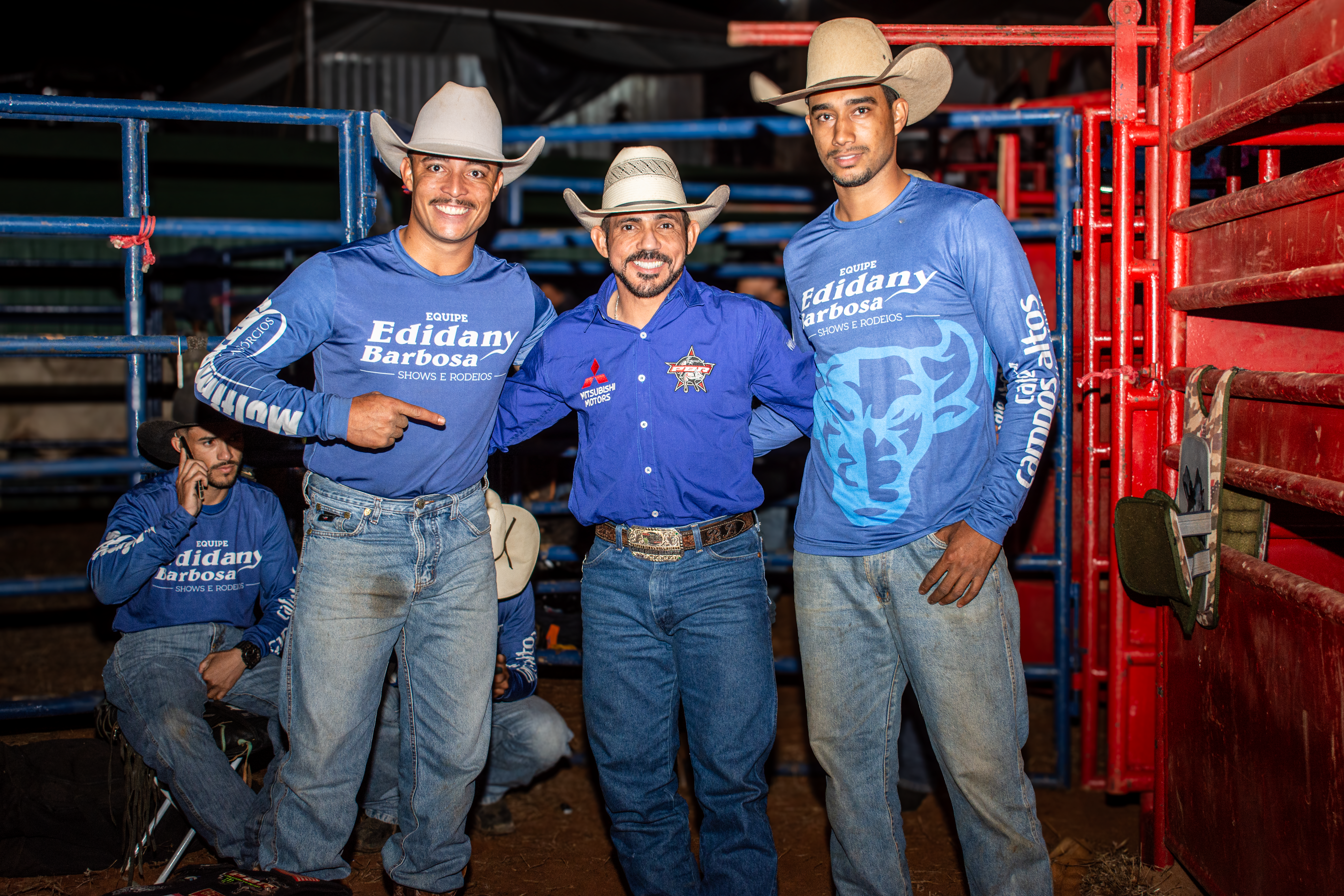 Three smiling cowboys pose at a rodeo in Tiros, Minas Gerais, Brazil, wearing cowboy hats and blue shirts.