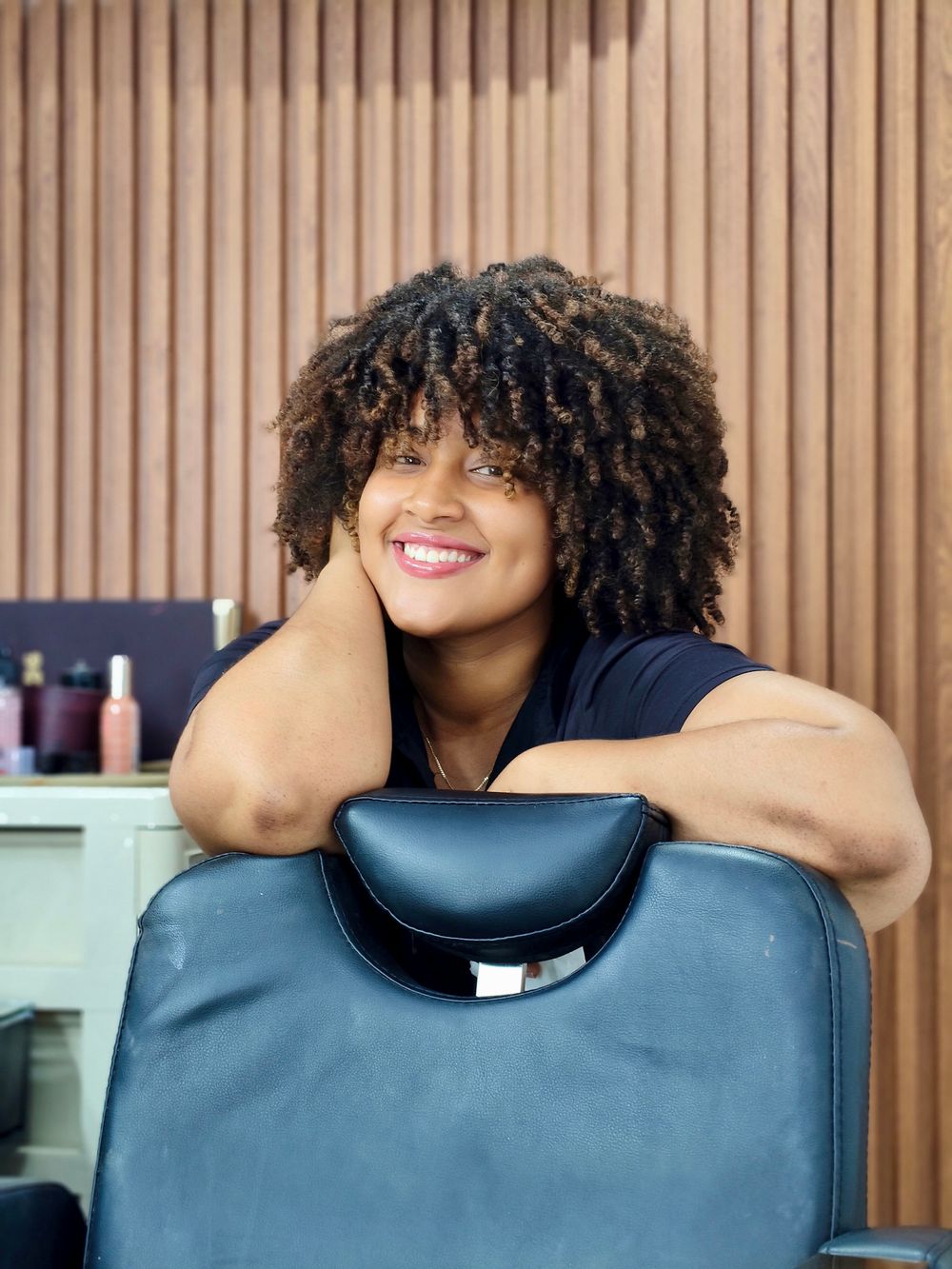 A smiling woman with defined curly hair leans on the back of a navy barber's chair against a wooden slatted wall.