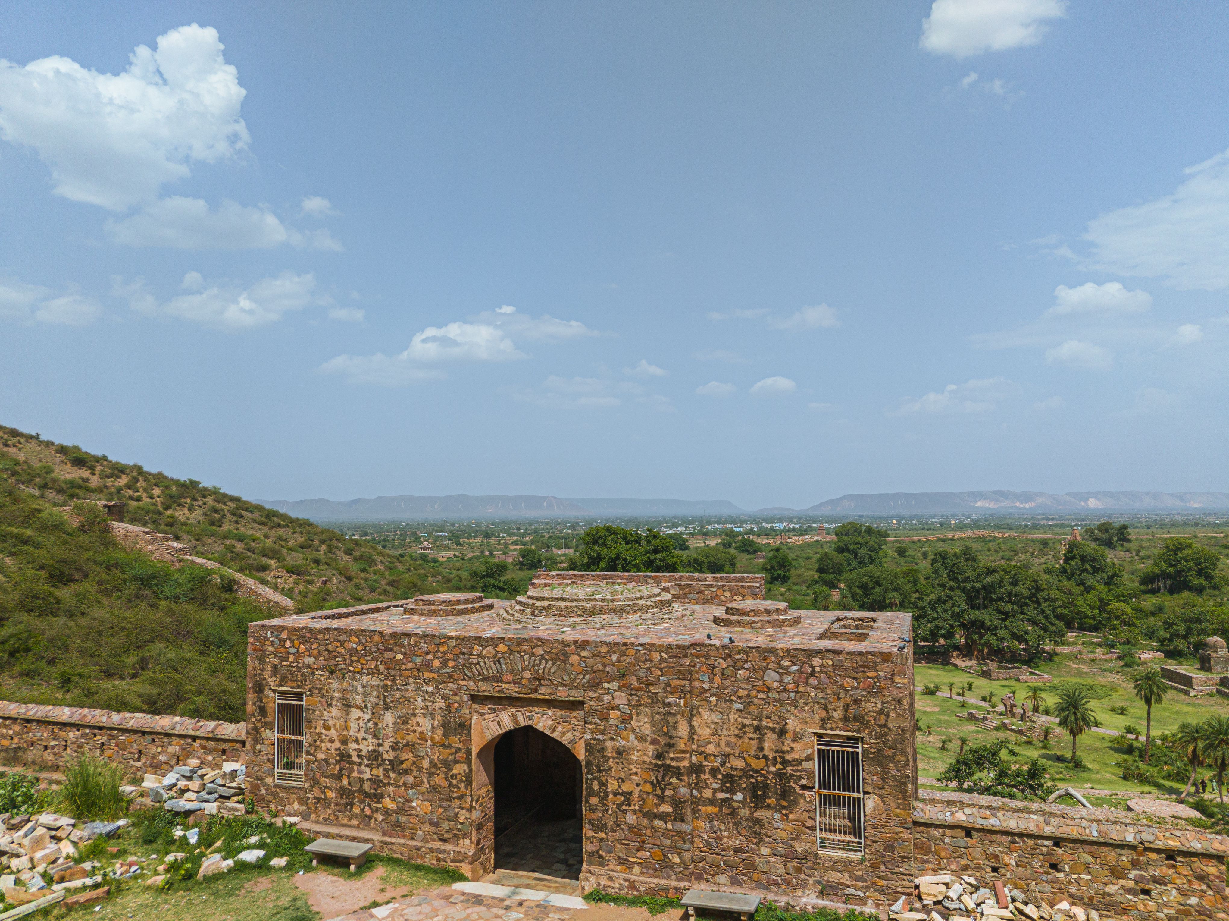 Ancient stone building amid rocky terrain and greenery under a bright sky.