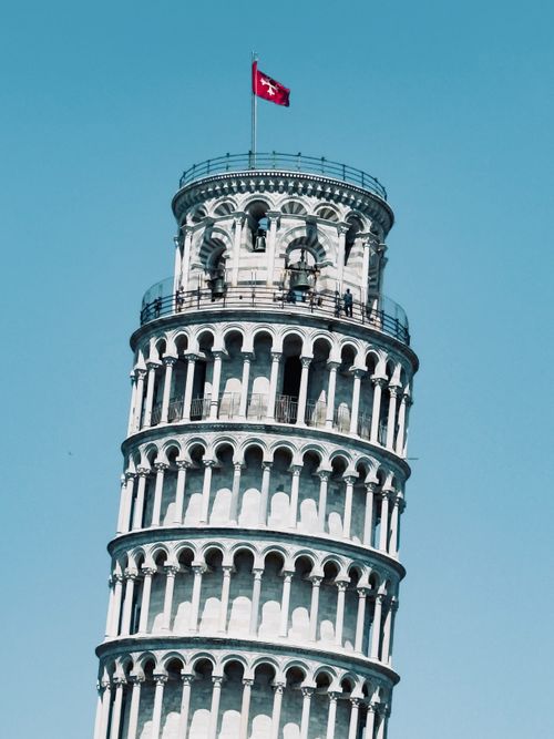 Free A striking view of the Leaning Tower of Pisa with a vibrant red flag, set against a clear blue sky. Stock Photo