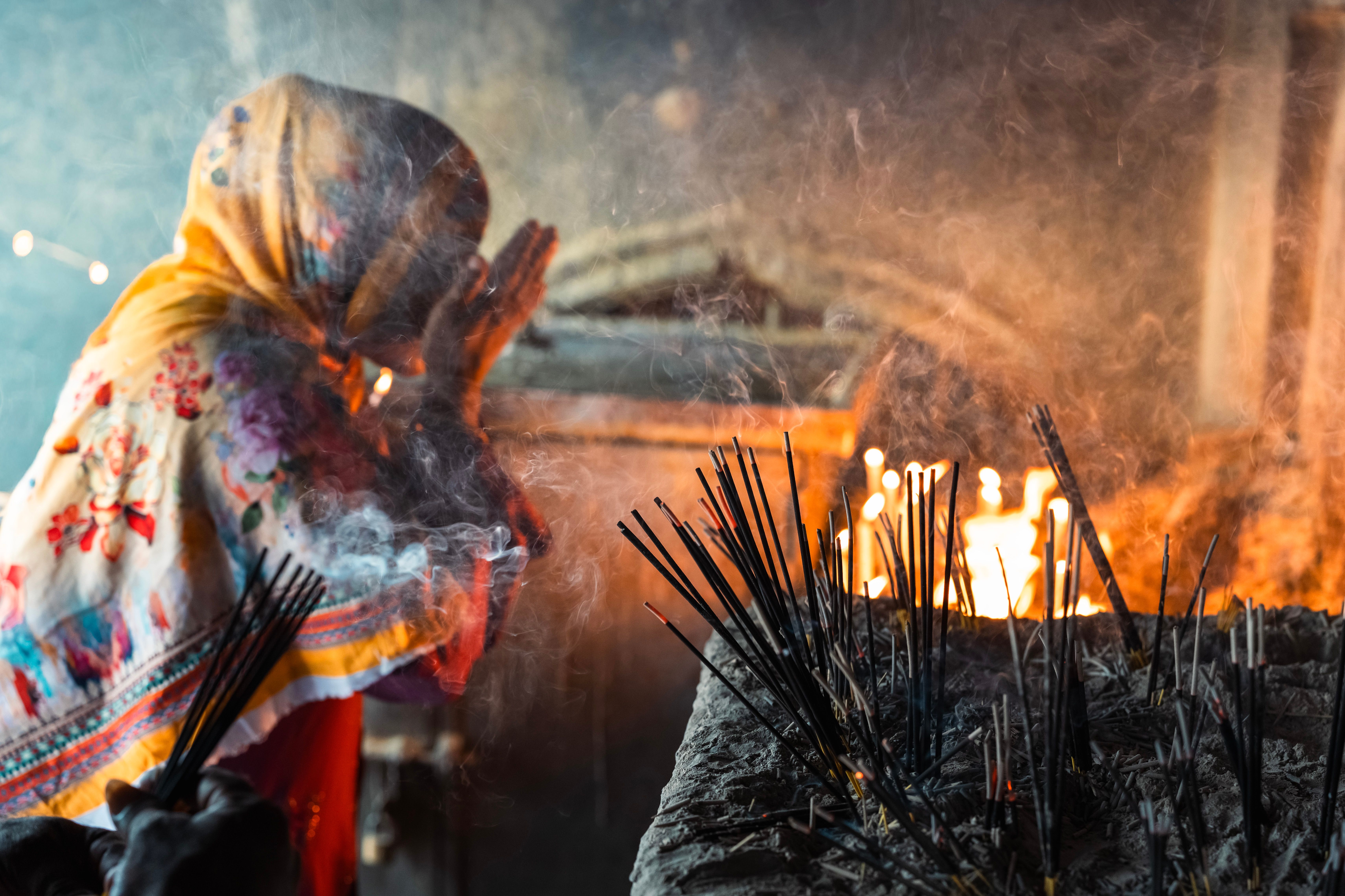 A woman in traditional attire praying with incense sticks in a dimly lit indoor setting.
