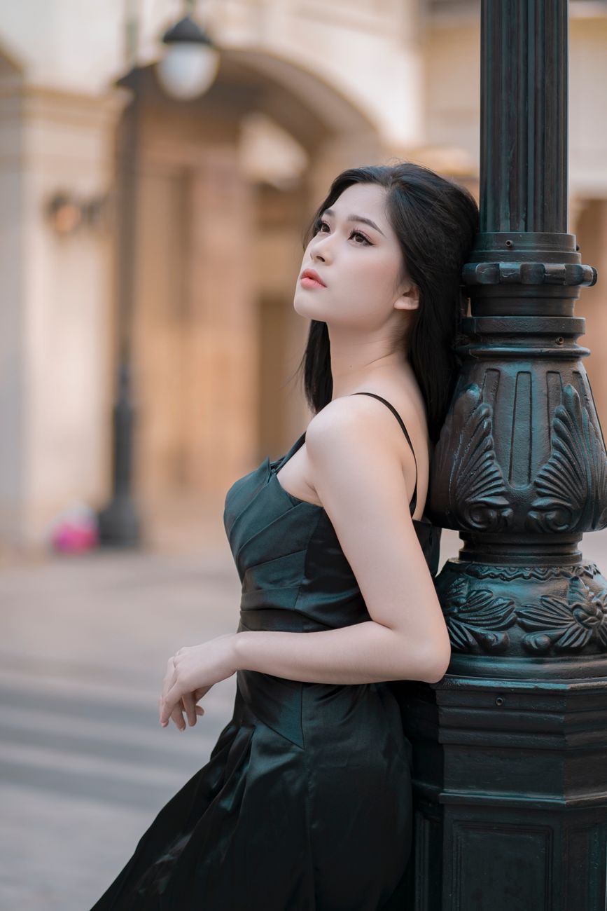 An elegant Asian woman posing outdoors in a stylish dress, leaning against a lamp post.
