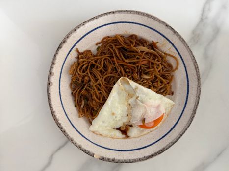 Top view of flavorful fried noodles with a fried egg on a rustic plate against a marble background.