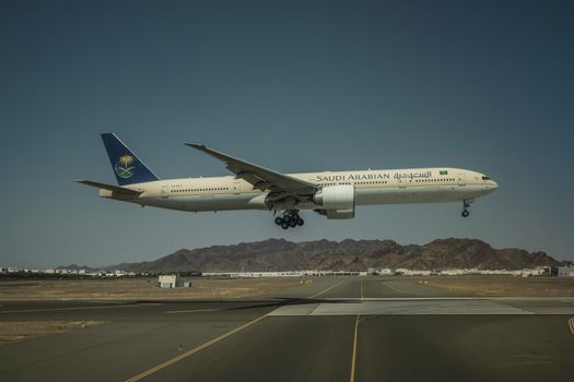 A Saudi Arabian Airlines Boeing 777 landing on a runway with mountainous backdrop.