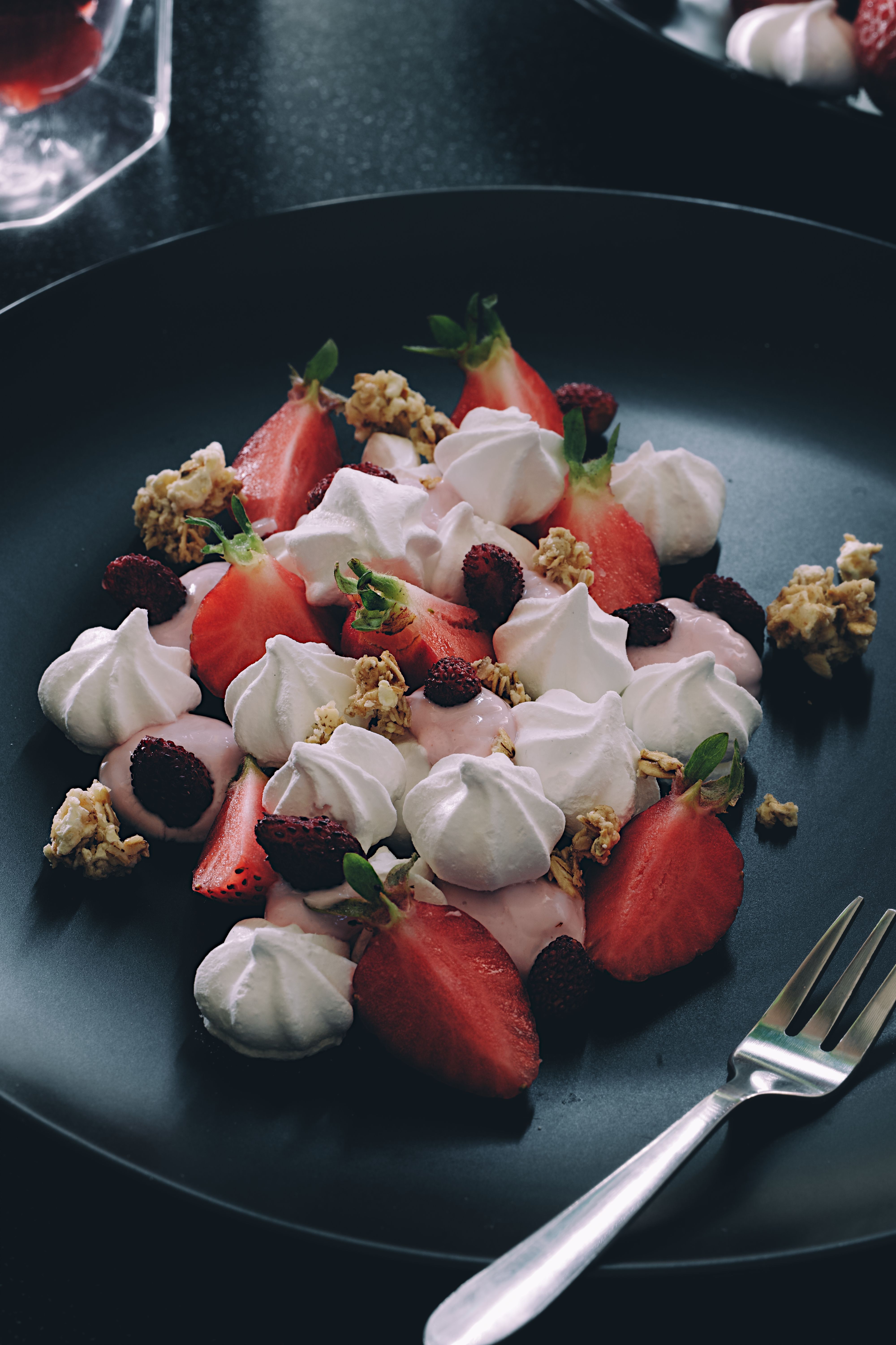 A mouthwatering dessert featuring strawberries, meringue, and granola on a dark plate.