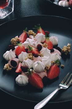 A mouthwatering dessert featuring strawberries, meringue, and granola on a dark plate.