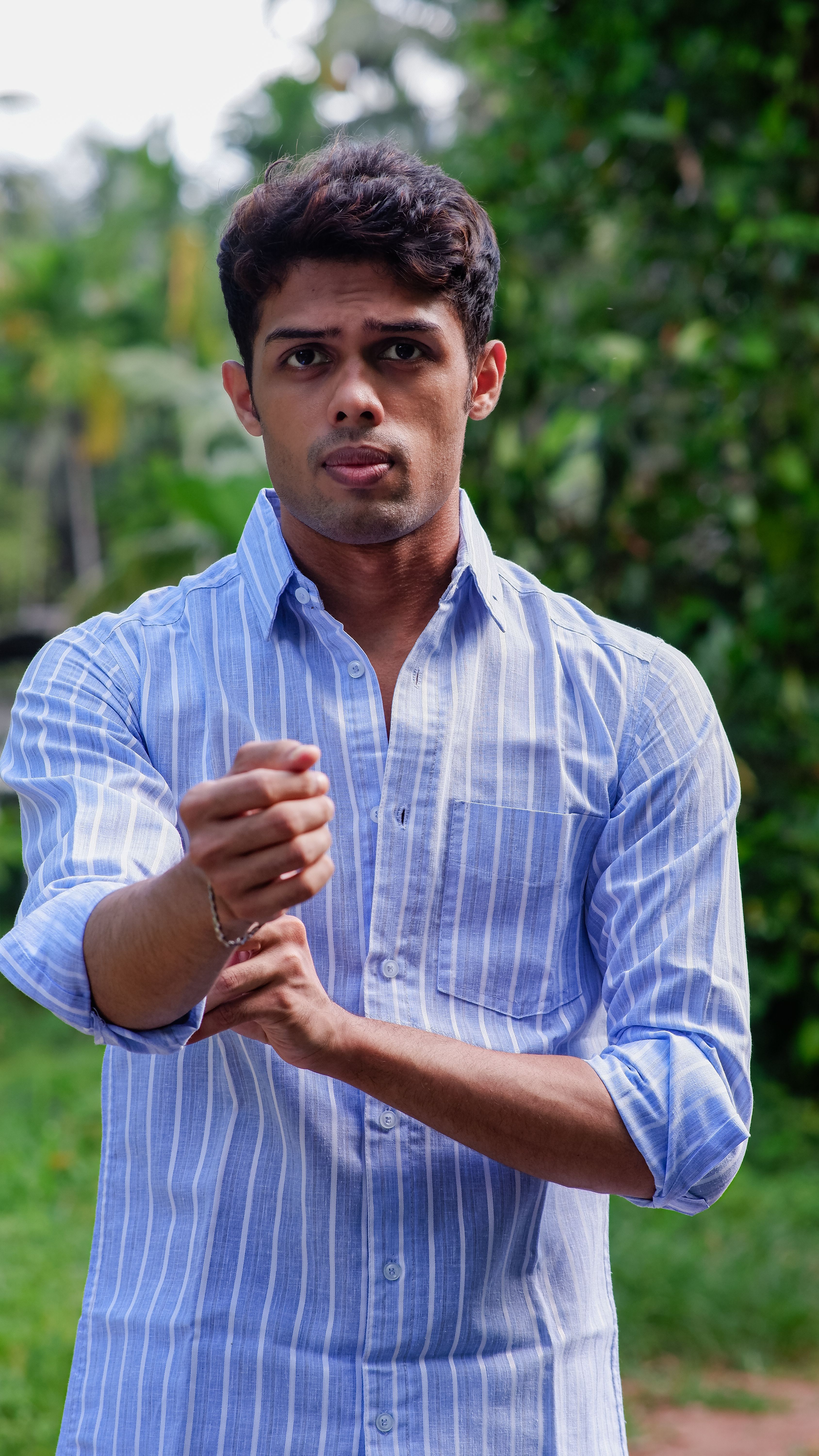 Portrait of a young man outdoors wearing a blue striped shirt, with a blurred natural background.