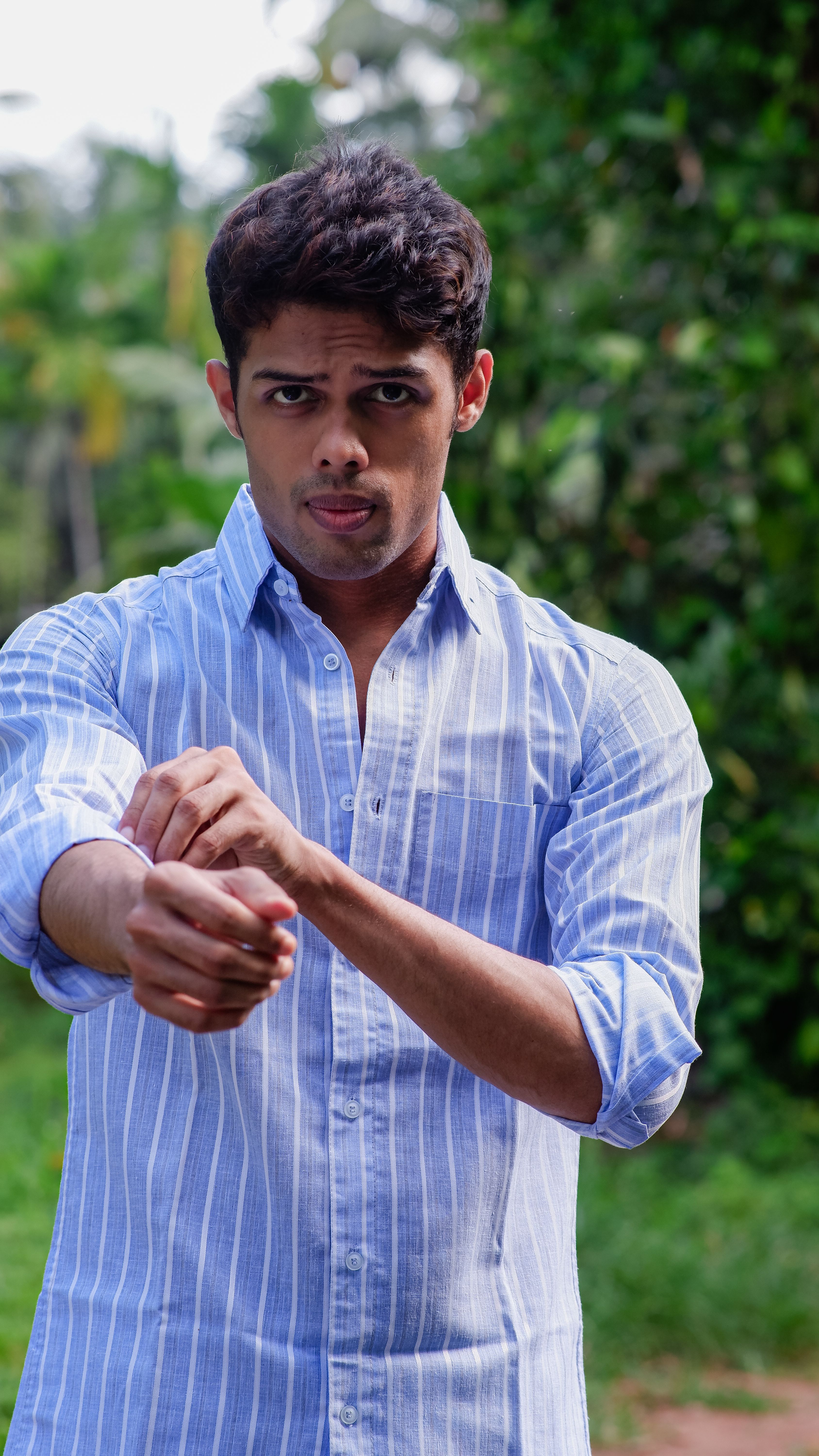 Confident young man with serious expression adjusting his sleeves in a natural outdoor setting.